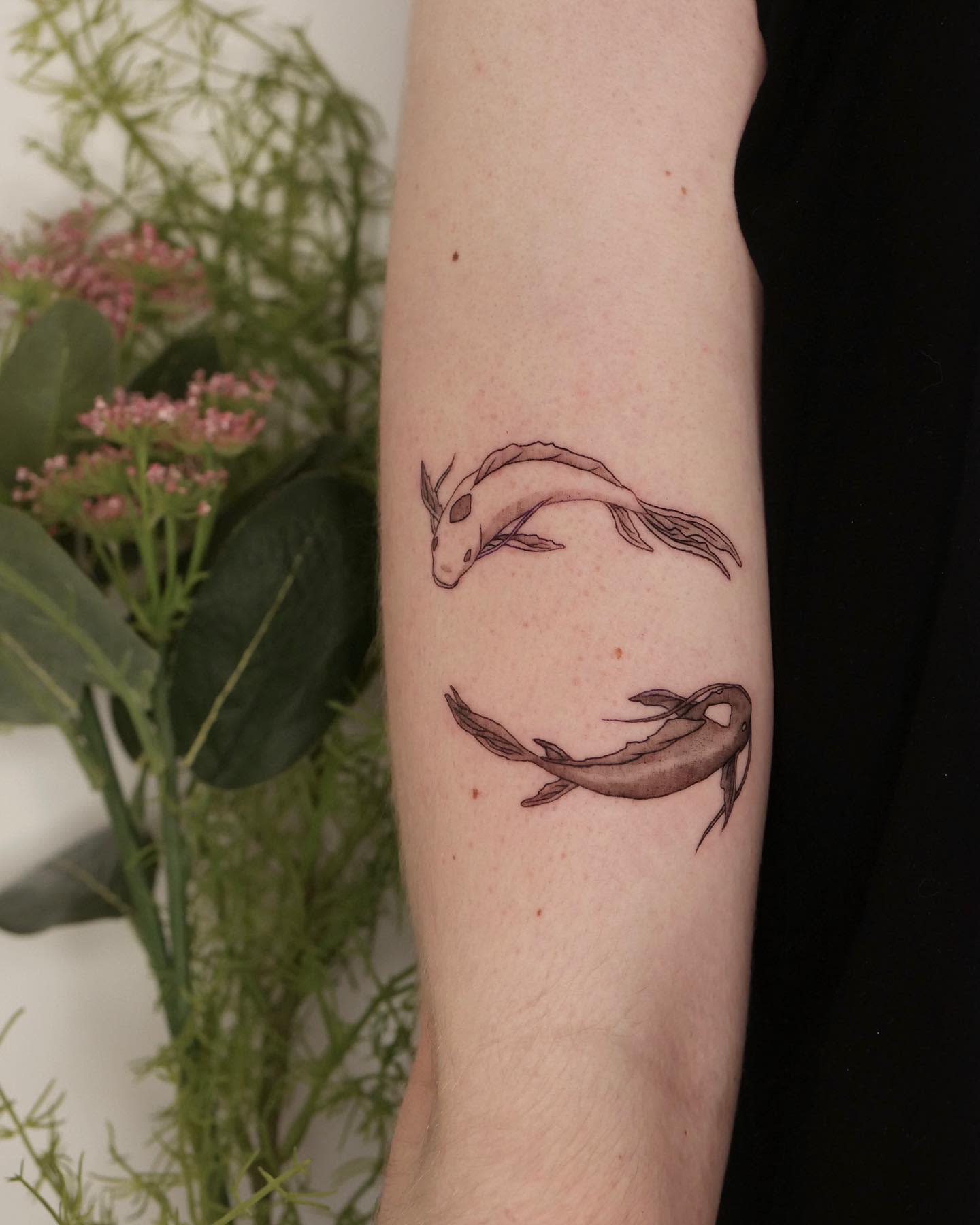 Two koi fish tattoos on an arm near plants, one black and the other light brown